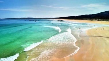 Havadan çekilen bir fotoğrafta, berrak turkuaz suları ve kıyıda hafif dalgaları olan güzel bir sahil sergileniyor.