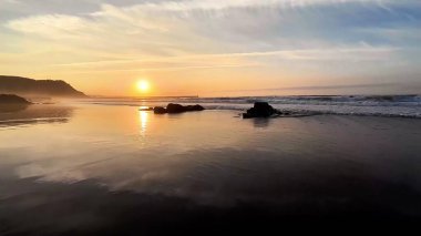 Sakin sahil sularında güzel renklerini yansıtan nefes kesici bir gün batımı. Nazik dalgalar kıyıya vururken.