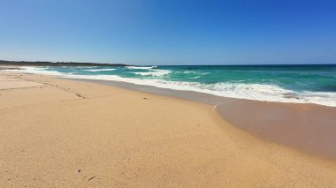 Huzurlu ve sakin bir sahil altın kum, berrak mavi gökyüzü ve yavaşça kıyıya vuran hafif dalgaları barındırır.
