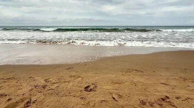 Yumuşak, altın kum, yumuşak dalgalar ve bulutlu gri gökyüzü içeren sakin ve sakin bir sahil manzarası.