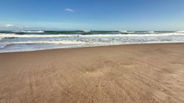 Yumuşak, sıcak kum, narin okyanus dalgaları ve yukarıda parlak, mavi bir gökyüzü sergilenen sakin bir sahil manzarası.