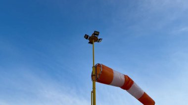 Şaşırtıcı derecede açık mavi bir gökyüzü, uzun bir direğe monte edilmiş çeşitli ışıklandırma ekipmanlarıyla birlikte renkli bir rüzgar çorabı sergiliyor.