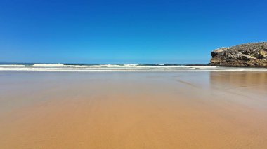 Huzurlu ve sakin bir sahil sahnesi. Dalgaların kıyıya vuruşunu ve yukarıda açık mavi bir gökyüzünü tasvir ediyor.