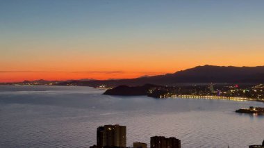 Canlı bir sahil şehrinin üzerinde, suda güzel yansıyan parıldayan ışıklarla nefes kesici bir günbatımı.