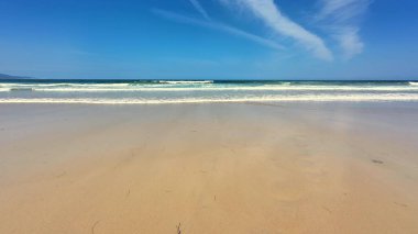 Sakin ve sakin bir plaj sahnesi yumuşak, sıcak kum, hafif dalgalar ve yukarıda açık mavi bir gökyüzü sergiliyor.