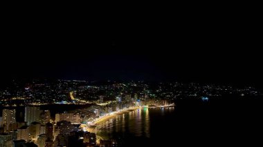 Gece boyunca kıyı boyunca sayısız parlak ışığın aydınlattığı canlı bir şehrin nefes kesici panoramik görüntüsü.