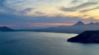 Dağların üzerinde çarpıcı bir günbatımı ve sakin bir deniz hayal gücünü cezbeden pitoresk bir sahil sahnesi yaratır.