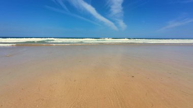 Huzurlu ve sakin bir sahil sahnesi yumuşak altın kumlar ve parlak mavi gökyüzünün altında yumuşak okyanus dalgaları.