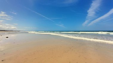 Yumuşak kum, yumuşak dalgalar, parlak mavi bir gökyüzünün yanında hoş ve huzurlu bir sahil sahnesi.