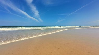 Huzurlu ve sakin bir sahil sahnesi yumuşak altın kum, berrak nazik dalgalar ve parlak mavi bir gökyüzü sergiliyor.