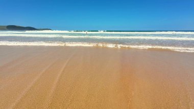Sakin bir sahil sahnesi, berrak mavi gökyüzü altında yumuşak dalgalar, rahatlamak ve gevşemek için mükemmel.