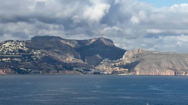 Nefes kesici bir manzara, dramatik bulutlu bir gökyüzü altında görkemli dağlar ve çarpıcı kıyı evleri barındırır.