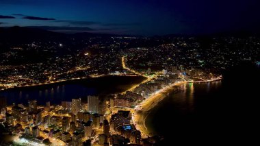 Göz kamaştırıcı, nefes kesici, canlı bir kıyı şehrinin gece gökyüzüne karşı güzelce aydınlatılmış görüntüsü.