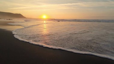 Çarpıcı bir günbatımı okyanus dalgaları üzerinde altın renkler bırakır. Huzurlu sahil atmosferini ve atmosferini yakalar.