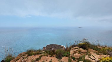 Güzel ve sakin bir kıyı manzarası kayalık kayalıklar, durgun masmavi sular ve bulutlu bir gökyüzü.