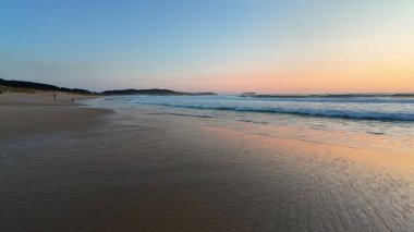 Gün batımında huzurlu ve sakin bir sahil sahnesi, hafif dalgalar ve gökyüzünde yayılan yumuşak renkler.