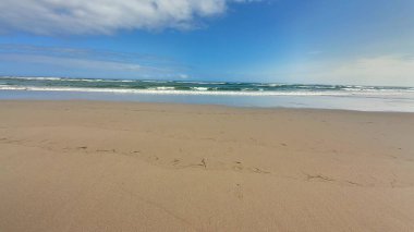 Görkemli bir plaj sahnesi. Altın kum, yumuşak dalgalar ve yukarıda parlak mavi bir gökyüzü.