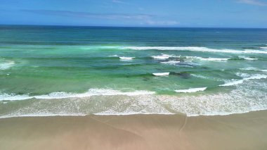Huzurlu bir sahil sahnesi yumuşak, nazik dalgaları sunar berrak, parlak mavi bir gökyüzünün altında kıyıya vurur
