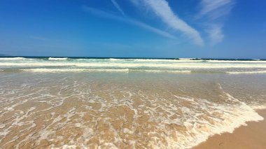 Nefes kesici bir manzara, açık mavi gökyüzünün altındaki kumlu kumsalda zarif dalgaları ortaya çıkarır.