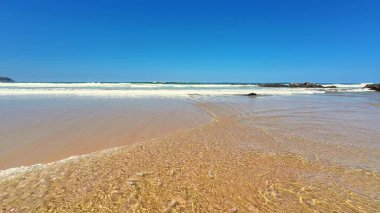 Huzurlu ve sakin bir sahil sahnesi yukardaki canlı mavi gökyüzü ile birlikte yumuşak, nazik dalgaları yakalıyor.