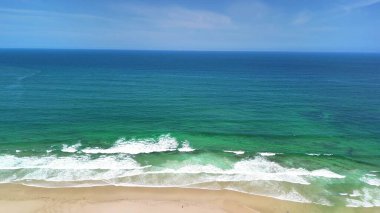Sakin bir sahilin havadan görünüşü yumuşak dalgaların kumlu sahili yalayıp yuttuğu açık mavi bir gökyüzü.