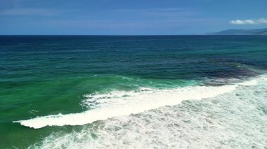 Hava görüntülerinde muhteşem turkuaz sular ve sakin dalgalar sergileniyor. Resimli kumlu bir plajda yavaşça yuvarlanıyorlar.