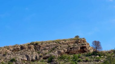 Sersemletici bir kayalık manzarası var, parlak yeşilliklerle süslenmiş, berrak mavi bir gökyüzünün altında.