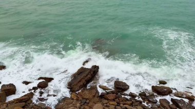 Dalgaların sakin suların ortasındaki engebeli kayaların üzerine çarptığı canlı bir sahil manzarası.