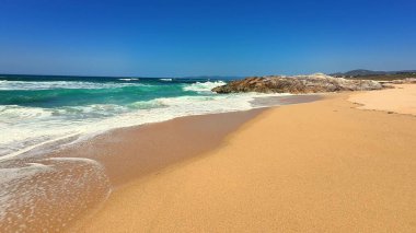Nefes kesen bir sahil manzarası, parlak, mavi gökyüzünün altında canlı dalgaların yanında altın kumlar sergiliyor.