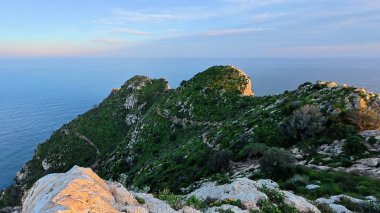 Serin denize bakan kayalık bir tepeden çarpıcı bir manzara, yemyeşil, canlı bitki örtüsüyle çevrili.