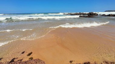 Sakin bir sahil sahnesi, parlak mavi gökyüzünün altında süzülen yumuşak dalgaları ve altın kumu gösteriyor.