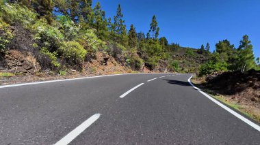 Dolambaçlı bir yol berrak mavi gökyüzünün altında parlak yeşilliklerle bezenmiş çarpıcı dağ manzarasının içinden geçiyor.