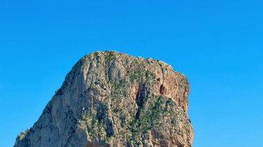 Bu nefes kesen çekim, dalgalı mavi gökyüzünün altında kristal berrak bir denizin üzerinde yükselen dik bir kaya yüzeyini yakalar.
