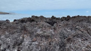 Kıyı şeridi boyunca uzanan engebeli kayaların çarpıcı manzarası bulutlu bir gökyüzünün altında güçlü dalgalar çarpıyor.