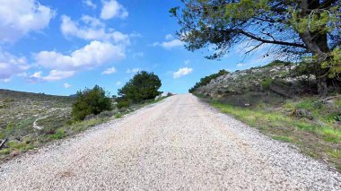 Güzel, manzaralı çakıllı bir yol, parlak, yemyeşil ve kayalık bir arazide, berrak mavi gökyüzünün altında yol alır.