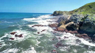 Dalgaların üstünden çekilen hava görüntüleri açık mavi gökyüzünün altında dramatik ve engebeli bir kayalık kıyıya çarpıyor.