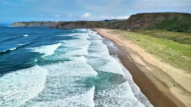 Uçurumlar ve yemyeşil yeşilliklerle çevrili kumlu bir plaja çarpan dalgaların nefes kesici görüntüsü..