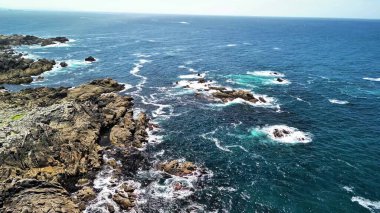 Turkuaz deniz dalgalarıyla birleşen kayalık bir sahil şeridinin çarpıcı hava görüntülerini görkemli bir manzara için tecrübe edin.