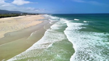 Sakin ve davetkar bir plaj sahnesi yumuşak dalgalar, güzel altın kum ve parlak mavi gökyüzü.