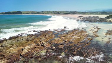 Nefes kesen güzellikte bir kıyı manzarası. Açık mavi gökyüzünün altındaki engebeli kayalara vuran dalgalar.