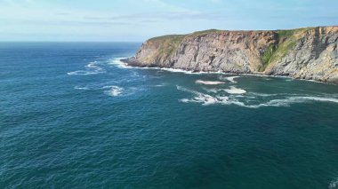 Açık mavi gökyüzünün altındaki dalgalar ve uçurumlarla kıyı manzarasını nefes kesici bir şekilde gözlemleyin.