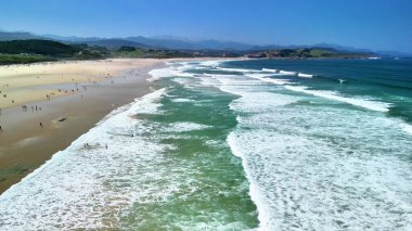 Çarpıcı bir hava manzarası dalgaların nazikçe çarptığı ve güneşlenenlerin mükemmel günün tadını çıkardığı canlı bir plajı sergiliyor.