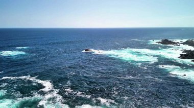 Açık mavi gökyüzünün altındaki kayalık bir sahile çarpan okyanus dalgalarının şaşırtıcı güzellikteki manzarasının tadını çıkar.