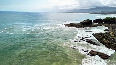 Okyanus kıyısındaki sarp kayalıkların çarpıcı manzarası güzelliği ve kıyı şeridini kaplayan nazik dalgaları vurguluyor.