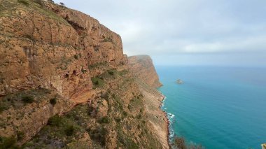 Bulutlu bir günde dingin mavi bir okyanusun yanında duran engebeli uçurumların nefes kesici manzarası.