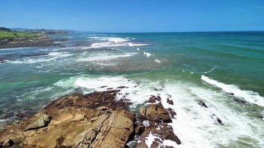 Görkemli okyanus dalgalarının çarpıcı bir görüntüsü berrak mavi gökyüzünün altındaki engebeli bir sahile çarpıyor.