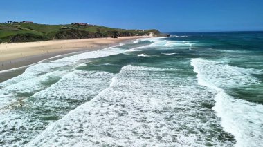 Güneşli bir günde altın kumlara çarpan canlı dalgaları canlı bir şekilde gösteren nefes kesici bir sahil manzarası.