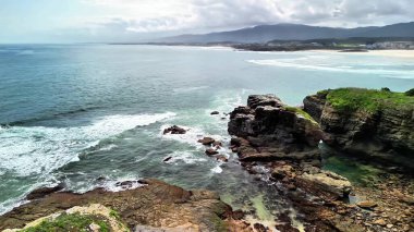 Çarpıcı kıyı manzarası görkemli dalgaları sergiliyor. Bulutlu bir gökyüzünün altında kayalık kıyılara şiddetle çarpıyor.
