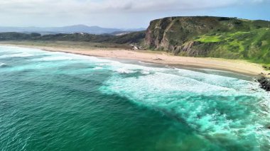 Okyanusun nefes kesici manzarası kumlu sahile çarpıyor, ve yemyeşil kayalıklar, hepsi berrak bir gökyüzünün altında