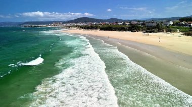 Bu, berrak mavi gökyüzünün altında yumuşak dalgalar ve altın kumlarla dolu canlı bir plajı gösteren çarpıcı bir hava manzarası.